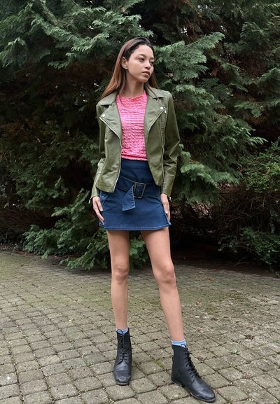 Chaqueta de cuero verde oliva, camisa texturizada rosa, falda mini de mezclilla azul con detalle de cinturón, botas de tobillo negras con cordones, en un entorno al aire libre.