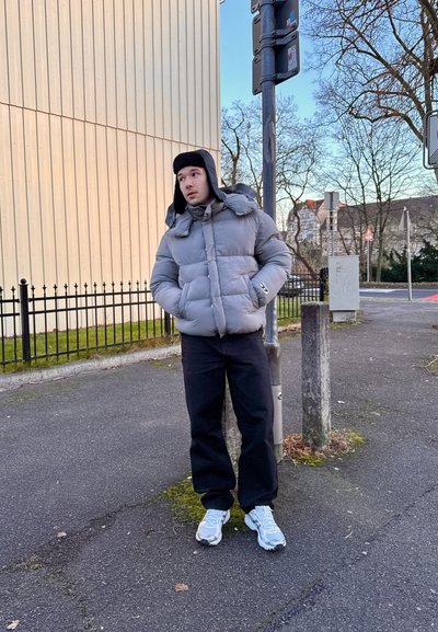 Chaqueta de plumas gris con capucha, con relleno grueso y un logo, pantalones negros y zapatillas de deporte azul claro. De pie junto a un poste en una acera.