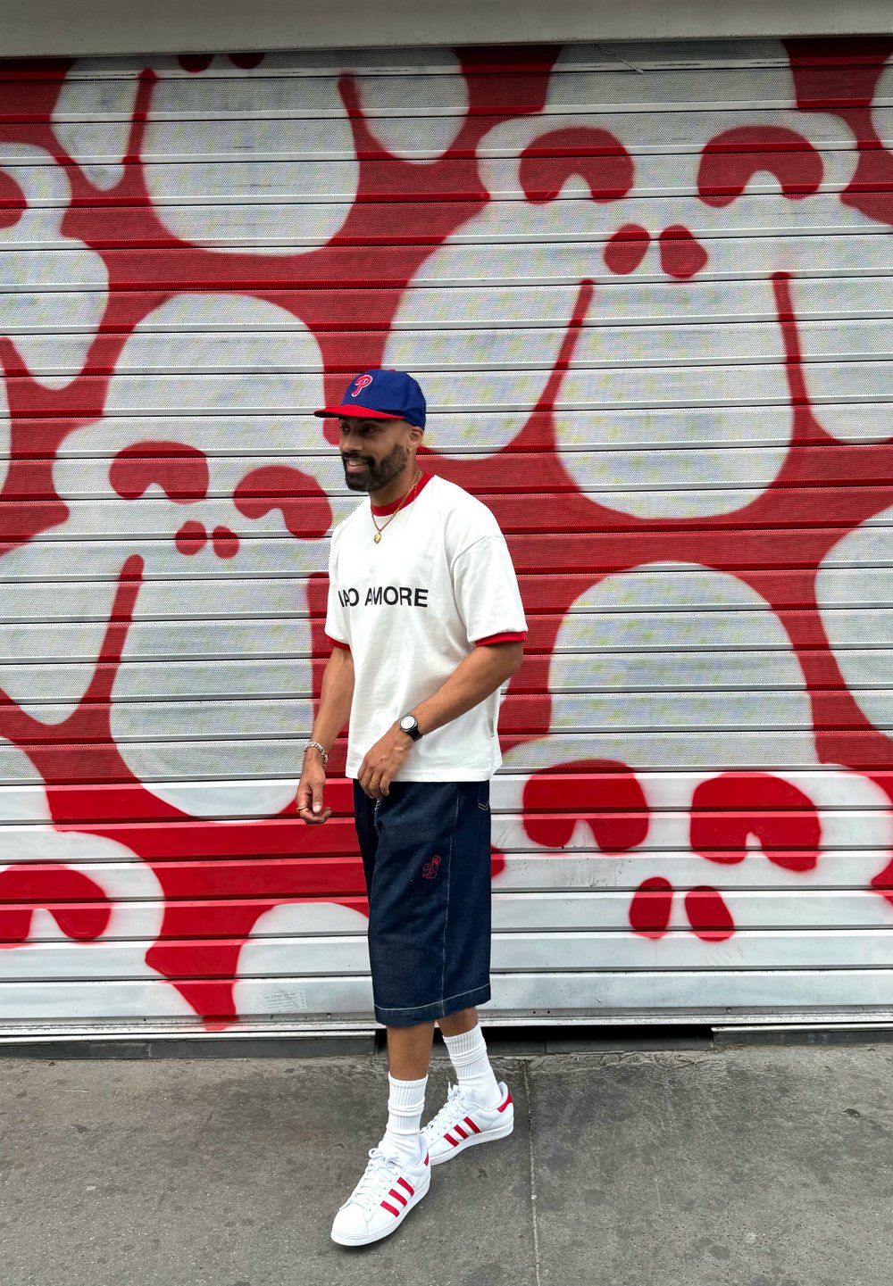 White t-shirt with "NO MORE" text, navy shorts, white sneakers with red accents, and a navy cap, walking past a red and white skull mural.