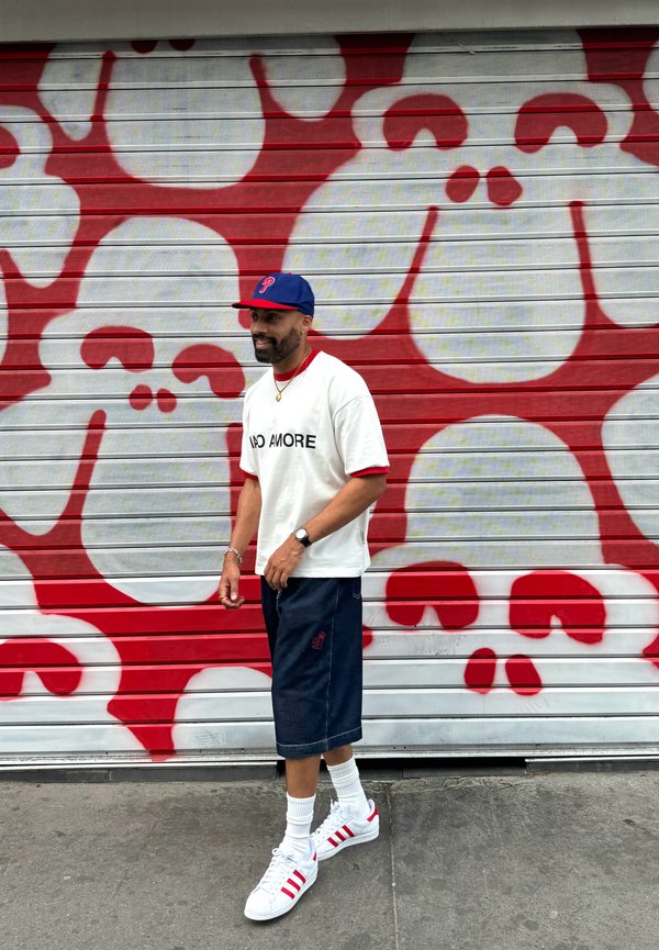 Hvit t-skjorte med teksten "NO MORE", marineblå shorts, hvite sneakers med røde detaljer, og en marineblå caps, går mot et rødt og hvitt hodeskallemurale.