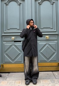 Black waterproof jacket with a hood, worn over a white shirt. Loose, faded grey trousers and black trainers. Background features green doors.
