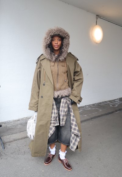 Mujer con un largo abrigo tipo gabardina color oliva, capucha forrada de piel, chaqueta beige, camisa de cuadros anudada a la cintura, falda vaquera, calcetines blancos y zapatos marrones, sosteniendo un bolso blanco.