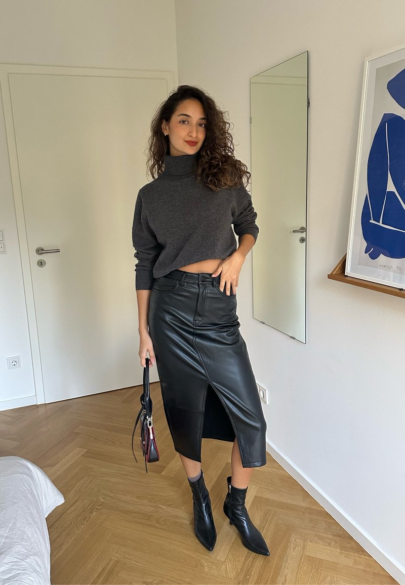 Grauer Rollkragenpullover, schwarze Leder-Bleistiftrock mit Schlitze vorne, schwarze Stiefeletten und eine kleine Handtasche, vor einem hellen Hintergrund.
