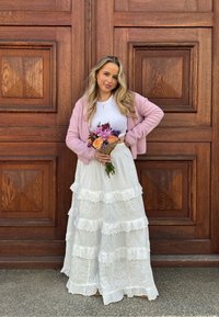 Helles rosa Strickjacke über einem weißen Oberteil, geschichteter weißer Rock mit Spitzenbesatz. Hält einen Blumenstrauß aus orangen und lila Blumen vor einem Holz-Hintergrund.
