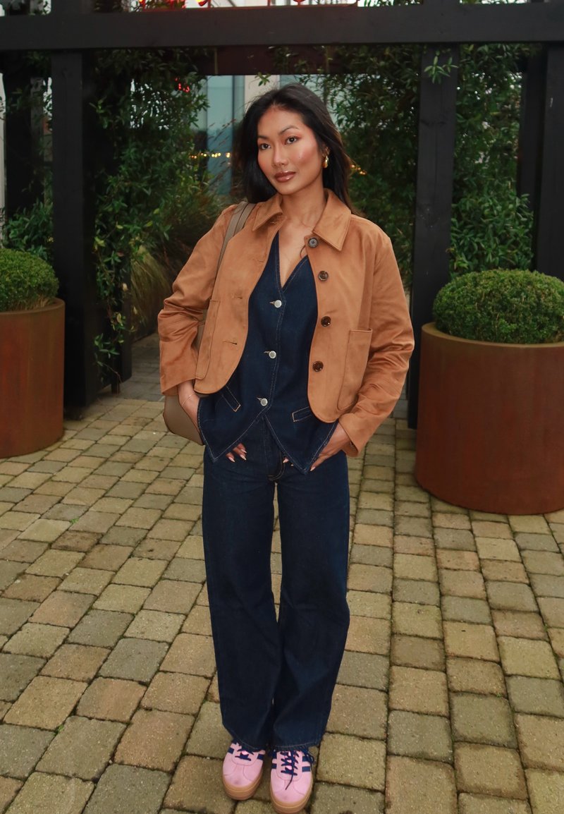 Brown suede jacket over a blue denim jumpsuit, light pink sneakers with navy laces, standing on a stone-paved surface with greenery in background.