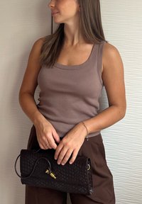 Zwarte geweven clutch met gouden hardware, vastgehouden in handen. Model draagt een aansluitend lichtbruin geribbeld tanktop en bruine broek. Glad achtergrond.