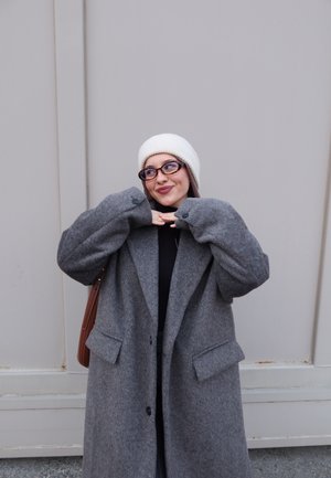 Joven con un abrigo gris oversize, gorro blanco de punto, gafas, sonriendo y posando con las manos bajo la barbilla contra un fondo liso.