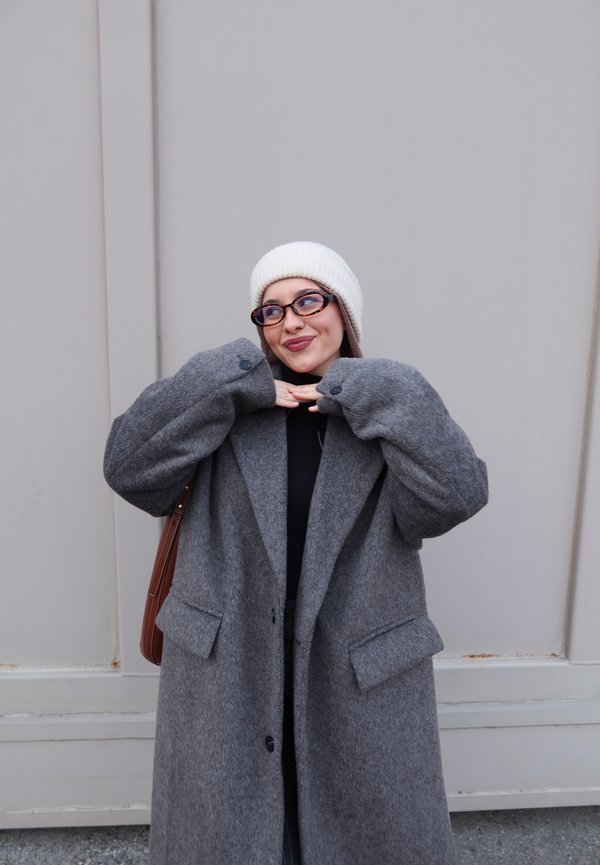 Jonge persoon die een grijze oversized jas draagt, een witte gebreide muts, een bril, glimlacht en poseert met de handen onder de kin tegen een effen achtergrond.