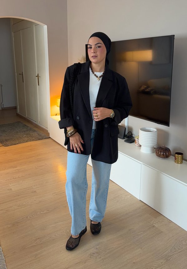 Mujer con pañuelo negro en la cabeza, blazer negro, camiseta blanca, vaqueros azul claro y zapatos negros, de pie en un salón moderno con suelo de madera.