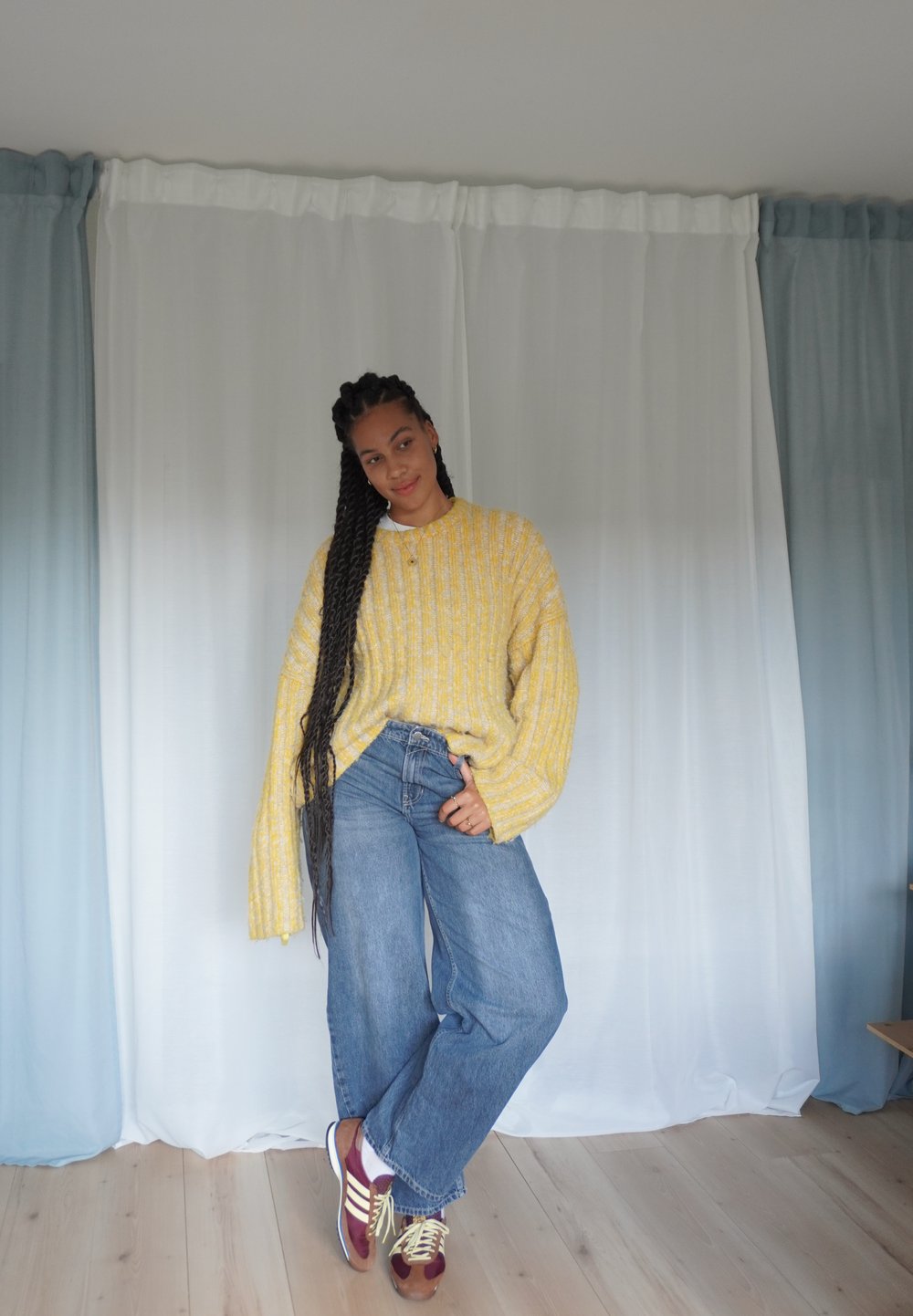 Pull en tricot jaune surdimensionné avec des rayures verticales, jean bleu à jambes larges et baskets bordeaux avec lacets jaunes, sur un fond clair.