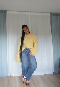 Pull en tricot jaune surdimensionné avec des rayures verticales, jean bleu à jambes larges et baskets bordeaux avec lacets jaunes, sur un fond clair.