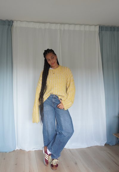 Suéter de punto amarillo de gran tamaño con rayas verticales, jeans de pierna ancha azul y zapatillas burdeos con cordones amarillos, todo sobre un fondo claro.