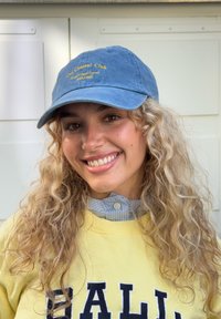 Giovane donna con capelli biondi ricci che indossa un berretto blu e una felpa gialla, sorridente in ambienti interni contro uno sfondo di parete bianca.