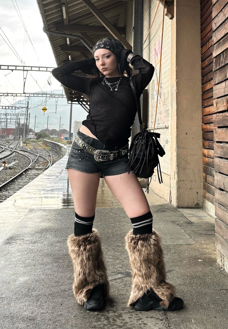 Jeune femme en haut noir, short, bottes en fourrure et bandana posant sur un quai de gare mouillé près des voies ferrées.