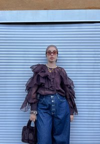 Top transparent de culoare mov închis cu mâneci volan, asortat cu blugi evazați albaștri cu talie înaltă. Accesoriile includ o poșetă neagră pufoasă și ochelari de soare.
