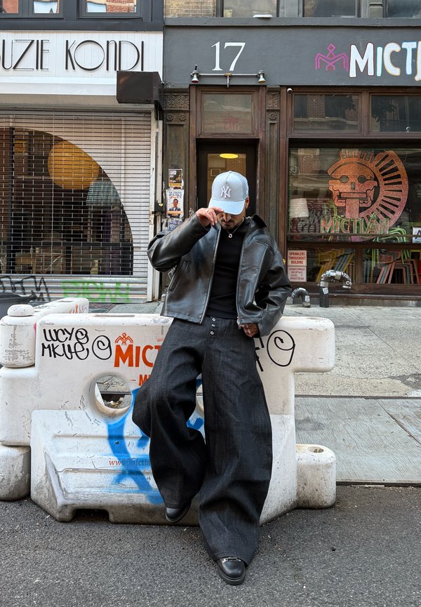 Black leather jacket, fitted black top, wide-leg dark denim trousers, grey cap with logo, standing against a graffiti-covered concrete barrier.