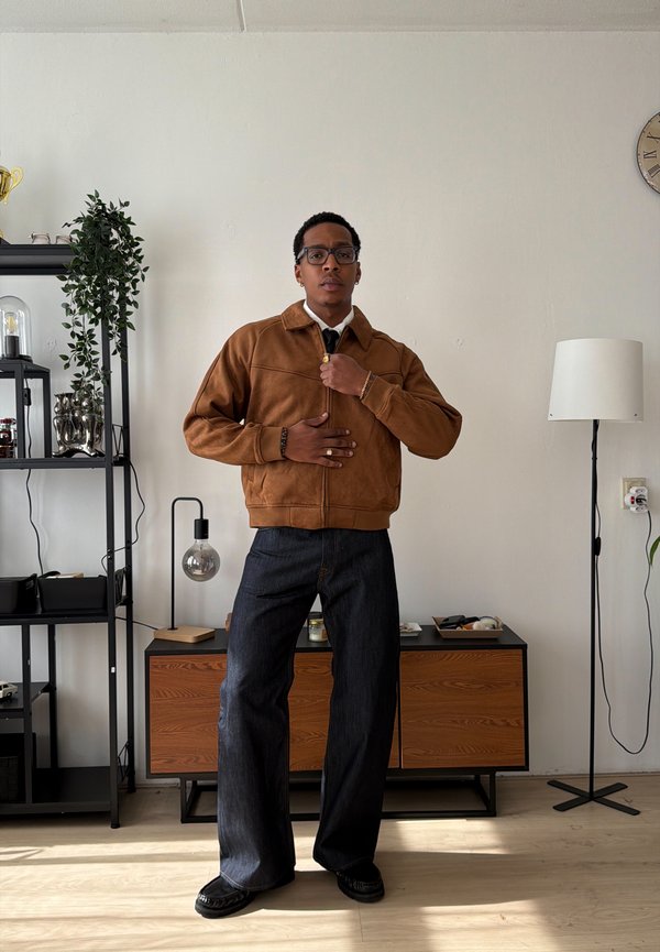 Man wearing brown jacket and dark wide-leg jeans standing in a room with shelves, a wooden cabinet, and a standing lamp.
