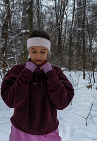 Sweat à capuche en polaire marron avec un logo blanc, superposé à une chemise violet clair. Un bandeau rose clair et un paysage hivernal avec de la neige.