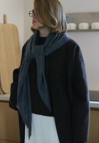 Siv volna šal okoli vratu; temen plašč in črni pulover spodaj; gladka tekstura; mehko padajoč; notranji prostor z lesenim ozadjem.