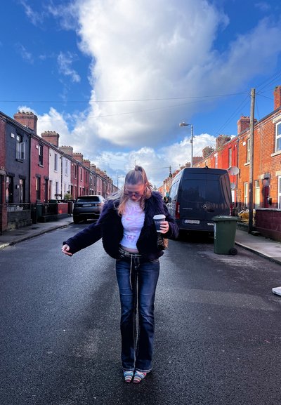 Joven con pelo largo, vestida con una chaqueta oscura y jeans, sosteniendo una taza de café, parada en una calle residencial con coches aparcados.