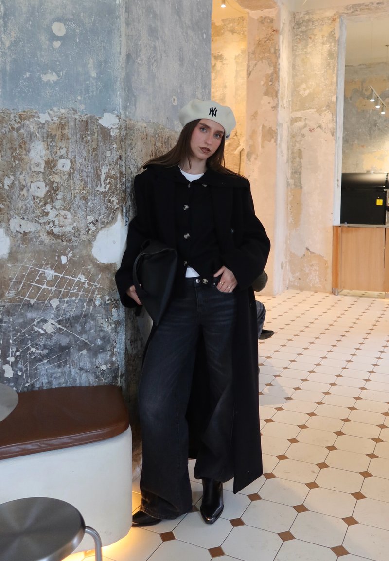Femme en manteau noir, pantalons larges et béret blanc appuyée contre un mur usé dans une pièce carrelée à la décoration vintage.