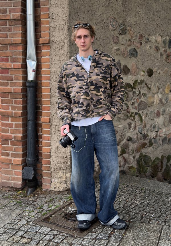 Junger Mann in einem Camouflage-Hoodie und weiten Jeans, der eine Kamera hält, steht auf einer Kopfsteinpflasterstraße neben einer Ziegel- und Steinmauer.