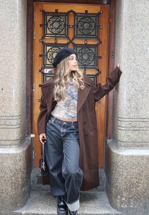 Mujer con boina negra, abrigo marrón, blusa estampada y vaqueros anchos, apoyada contra pilares de piedra y una puerta de madera con rejas de metal ornamentadas.