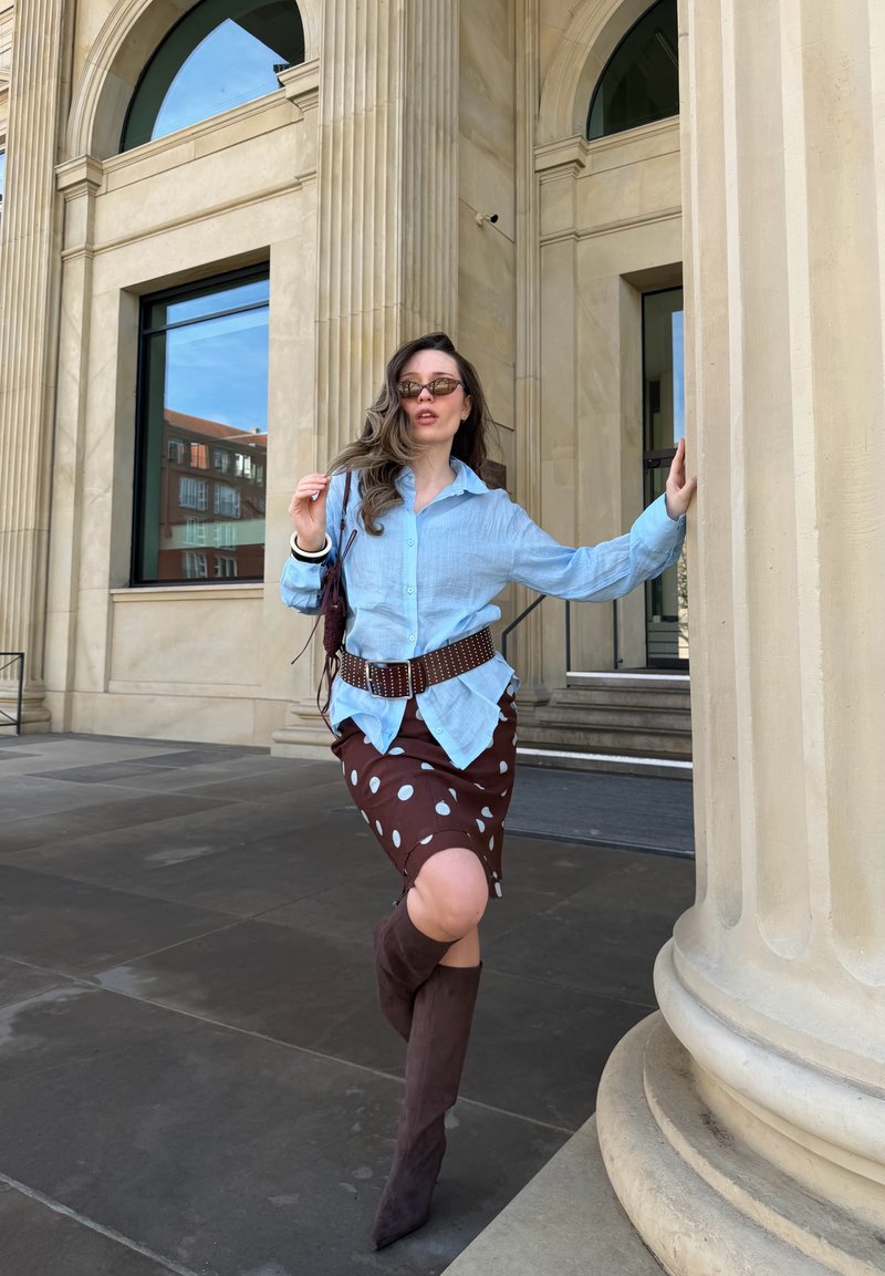 Femme posant à côté d'une grande colonne, portant une chemise bleu clair, une jupe marron à pois, des bottes marron, des lunettes de soleil et tenant un petit sac.