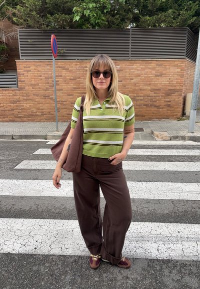 Mujer con camiseta de rayas verdes, pantalones marrones y gafas de sol, de pie en un paso de peatones con un bolso marrón al hombro.