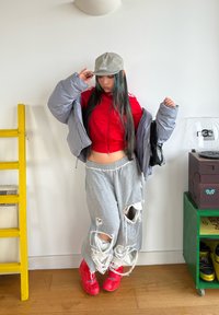 Punainen lyhyt takki, ylipitkä harmaa untuvatakki, repeytyneet harmaat collegehousut ja kirkkaan punaiset kengät. Hatussa Adidas-logo. Huoneen tausta.