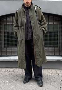 Homme portant un long trench-coat vert olive, une chemise noire, un pantalon foncé et des chaussures noires, debout sur un trottoir carrelé devant des fenêtres grillagées.
