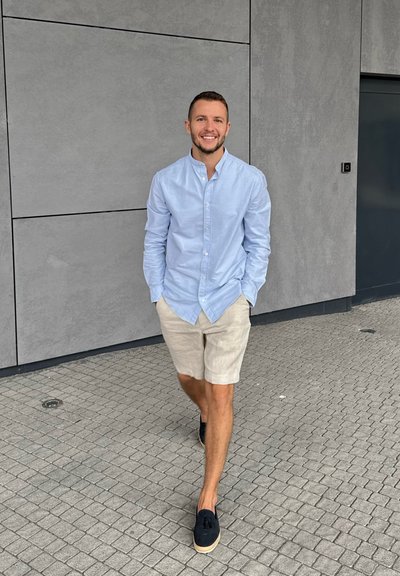 Camisa de botón de color azul claro, pantalones cortos beige y zapatos oscuros. Materiales suaves, corte relajado, caminando sobre una superficie pavimentada contra una pared gris.