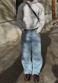 Felpa grigia, jeans a gamba larga blu chiaro e scarpe in pelle verniciata scura. Sfondo stradale con texture e ombre create dalla vegetazione vicina.
