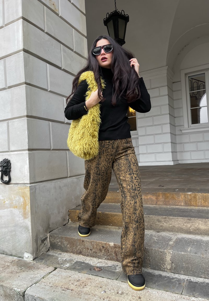 Haut en maille noir, sac à épaule jaune en fourrure, pantalon large à motif serpent marron, et chaussures noires avec semelles jaunes, debout sur des marches en pierre.