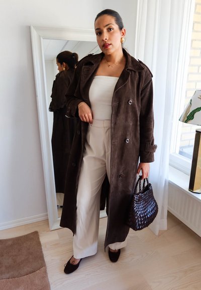 Mujer con abrigo largo marrón, camiseta blanca y pantalones anchos beige que sostiene un bolso oscuro tejido, de pie frente a un espejo en una habitación luminosa.