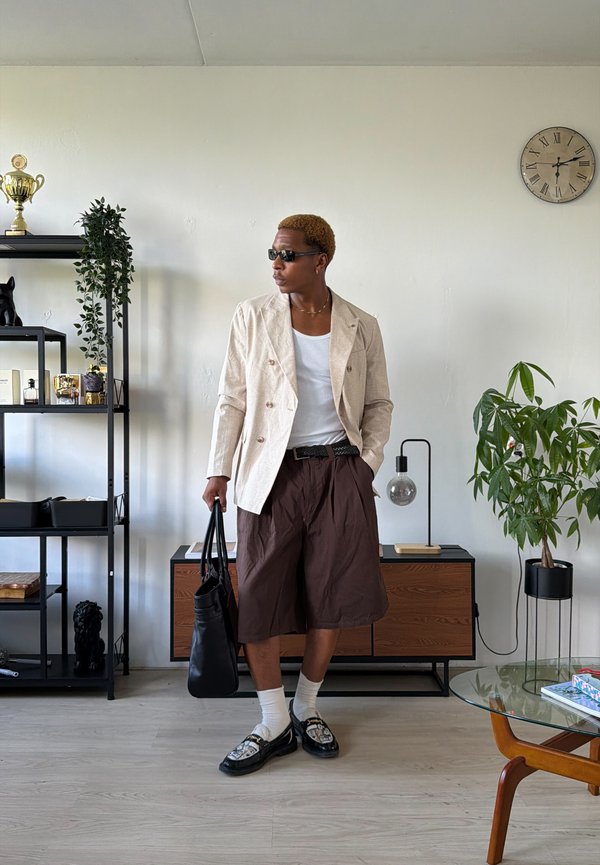 Hombre con gafas de sol que lleva una chaqueta beige, camisa blanca y pantalones cortos marrones, está de pie sosteniendo una bolsa negra en una sala de estar moderna con plantas y un reloj.