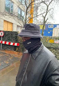 Svart buckethat med Y-3-logga, svart ansiktsskydd och en svart regnjacka med en glänsande yta, stående i en regnig urban miljö.