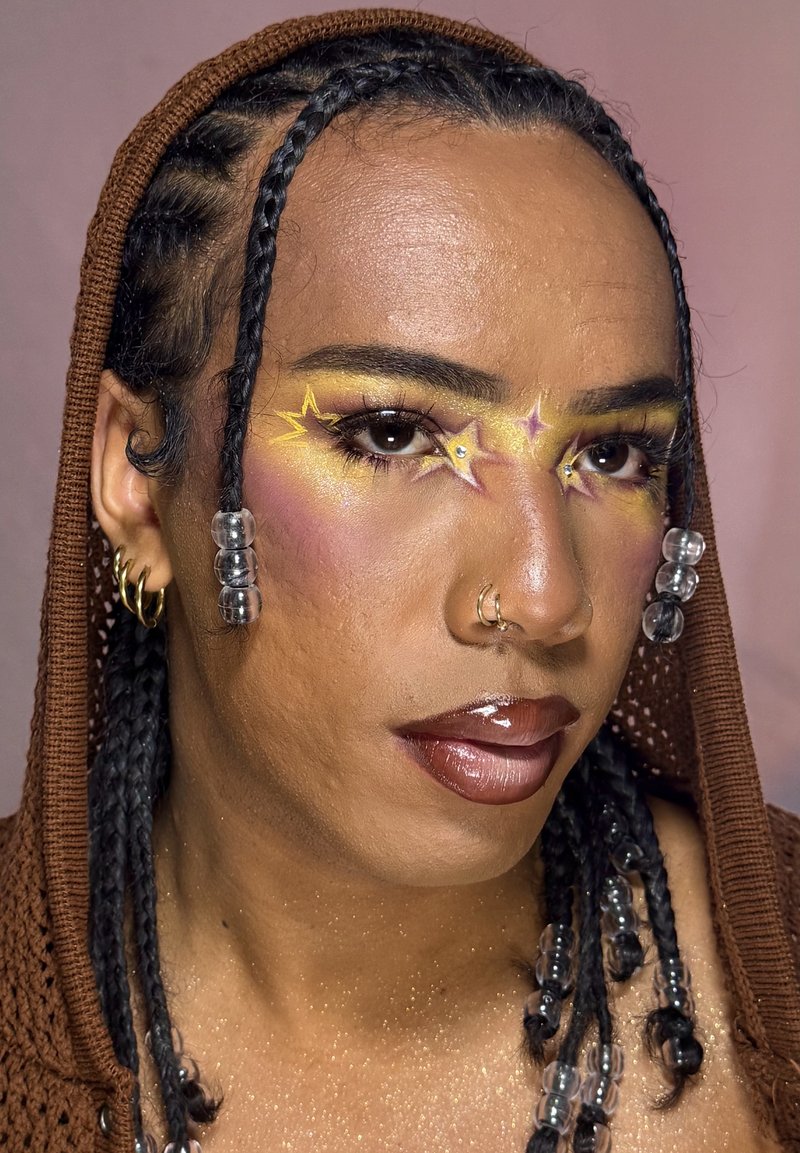 Person mit geflochtenem Haar, das mit klaren Perlen verziert ist, gelbem Stern- und Glitzerschmuck im Gesicht, glänzenden dunklen Lippen, Nasenpiercing, goldenen Creolen und braunem Kapuzenpullover.