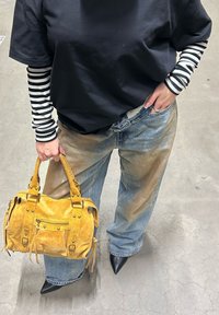Sac à main en cuir jaune avec des clous métalliques, tenu dans une main. Haut noir à manches courtes porté sur un chemisier à manches longues rayé noir et blanc, jean bleu ample, et escarpins noirs pointus.