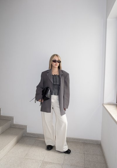 Mujer con blazer gris de gran tamaño, top negro, pantalones blancos de pierna ancha, zapatos negros, gafas de sol, sujetando bolso negro, de pie junto a unas escaleras y una ventana.