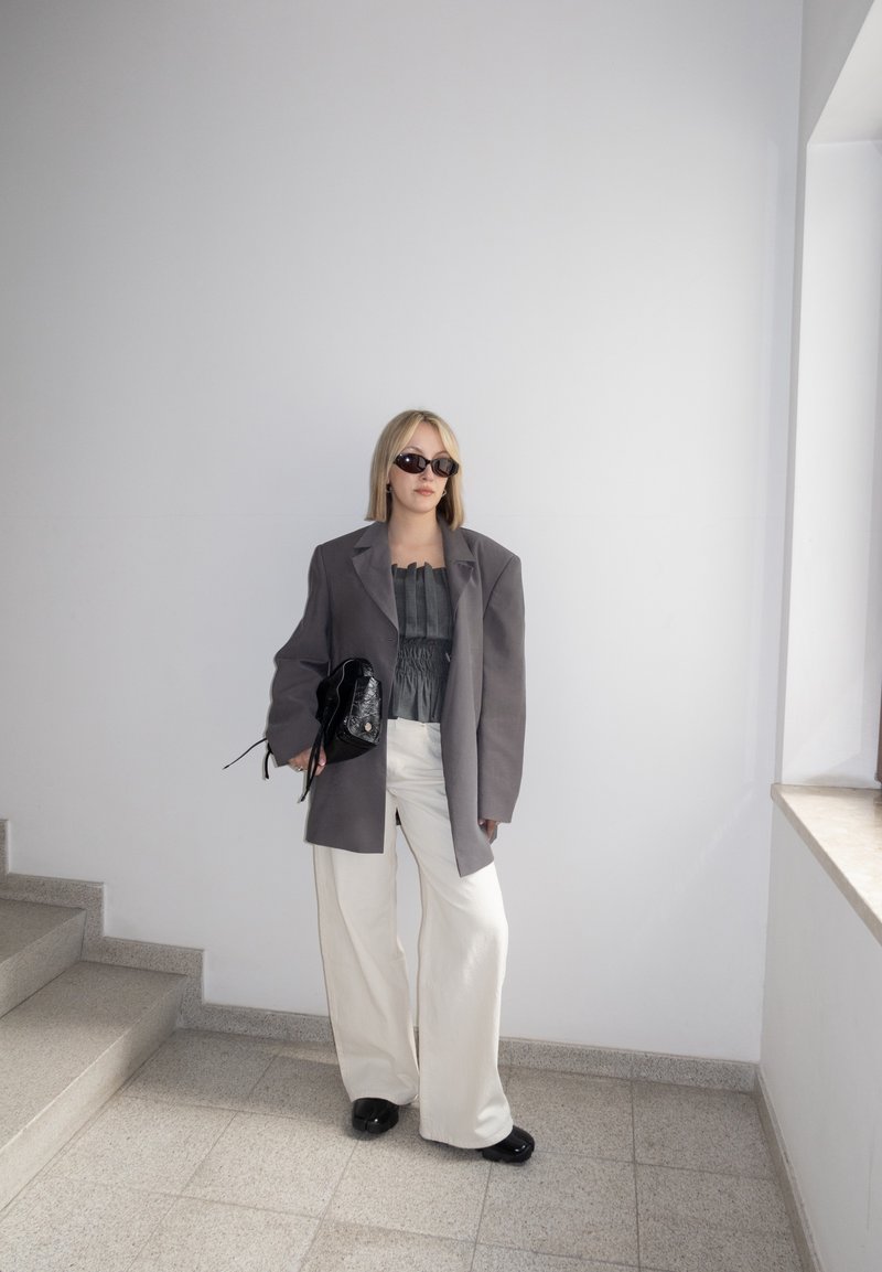 Femme portant un blazer gris surdimensionné, un haut noir, un pantalon blanc à jambes larges, des chaussures noires, des lunettes de soleil, tenant un sac noir, debout près des escaliers et d'une fenêtre.