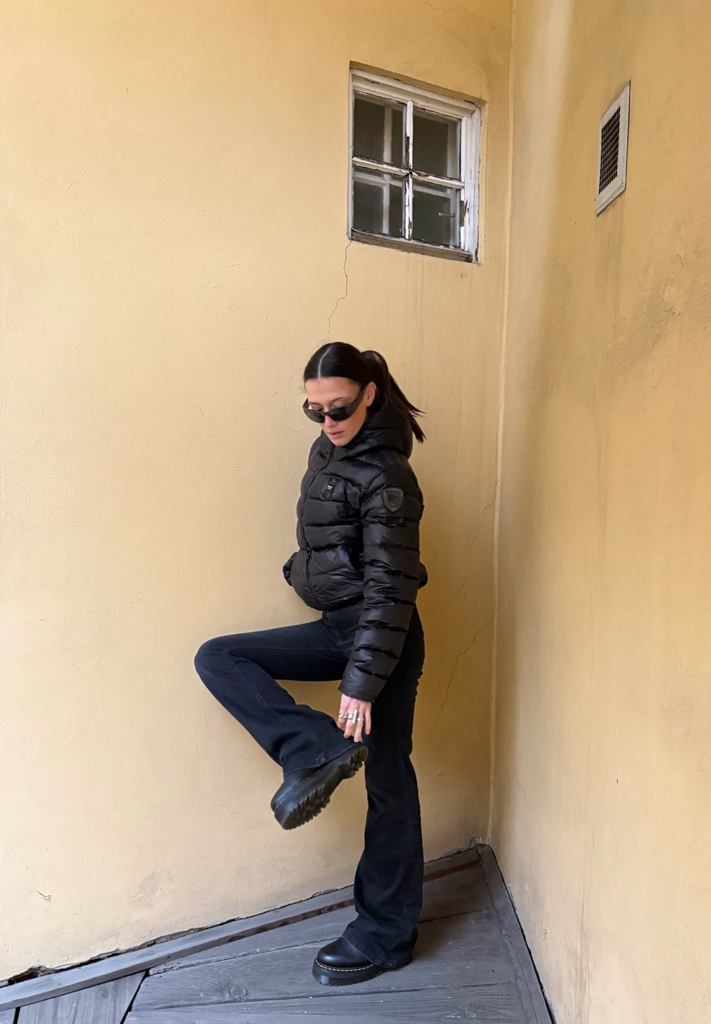 Veste matelassée noire avec un design matelassé horizontal, associée à un jean évasé foncé et des bottes à plateforme épaisses noires. Fond neutre.