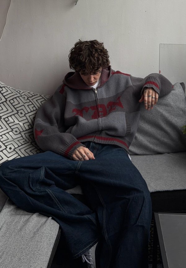 Lockighaarige Person in einem übergroßen grauen und roten Pullover und weiten blauen Jeans sitzt auf einem Sofa mit gemusterten und einfarbigen Kissen und schaut nach unten.