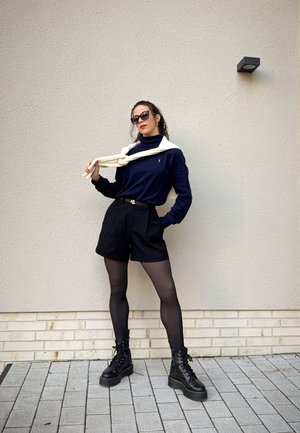 Suéter de cuello alto azul marino, pantalones cortos negros de cintura alta, medias negras y botas de combate. Suéter ligero sobre los hombros. Fondo minimalista.