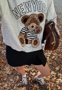 Hanorac gri cu broderie cu urs, pantaloni scurți negri, pantofi sport maro și o geantă maro. Așezat printre frunze căzute.