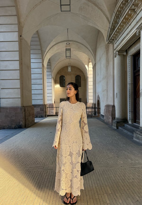 Mujer con un largo vestido beige de encaje que sostiene un bolso negro, está de pie en un pasillo de piedra con arcos y techos abovedados iluminado por el sol.