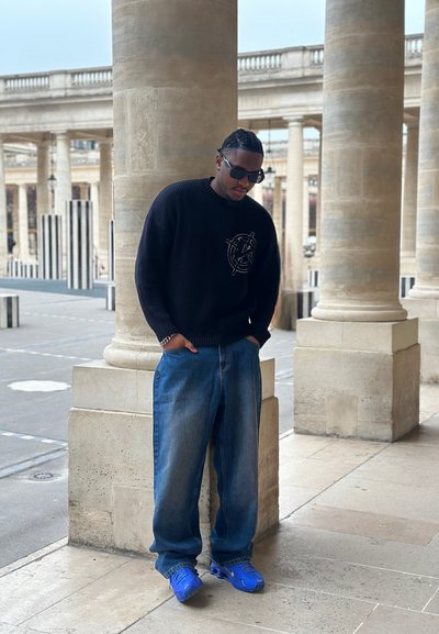 Hombre con suéter negro, jeans azules y zapatillas azul brillante, de pie con las manos en los bolsillos cerca de columnas de piedra al aire libre.
