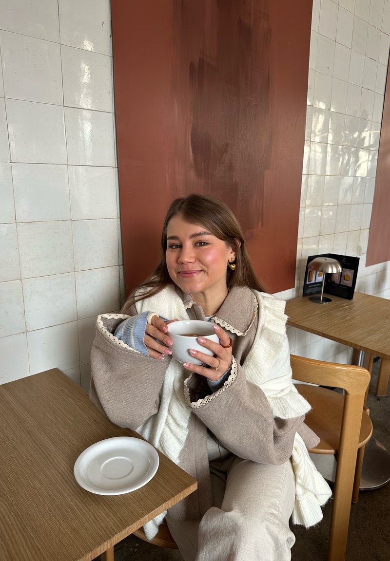 Jeune femme vêtue de beige et blanc, assise à une table de café, tenant une tasse blanche à deux mains, souriant doucement.