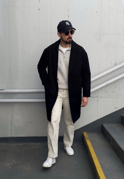 Abrigo negro sobre un suéter ligero, pantalones crema y zapatillas blancas. Los accesorios incluyen una gorra negra y gafas de sol. El fondo presenta concreto y escaleras.
