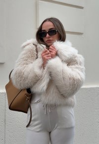 Κρεμ ψεύτικη γούνα ζακέτα με cropped σχέδιο και μεγάλο γιακά. Συνοδεύεται από καφέ τσάντα και ανοιχτό γκρι παντελόνια φόρμας. Τα γυαλιά ηλίου ολοκληρώνουν την εμφάνιση.